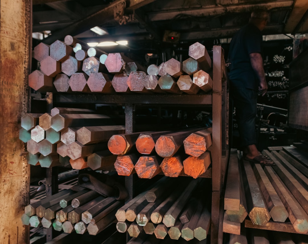 An industrial stockyard, featuring stacked hexagonal and circular metal bars on shelves. The varied ends of the bars display a spectrum of colors—from rust-red to oxidized green—creating a dense, repetitive pattern and geometric complexity. A worker on the right adds a sense of human scale to the heavy inventory. This texture is highly useful for engineering firm advertisements, as a conceptual background representing raw material wealth, or in editorial content about manufacturing and global supply chains.